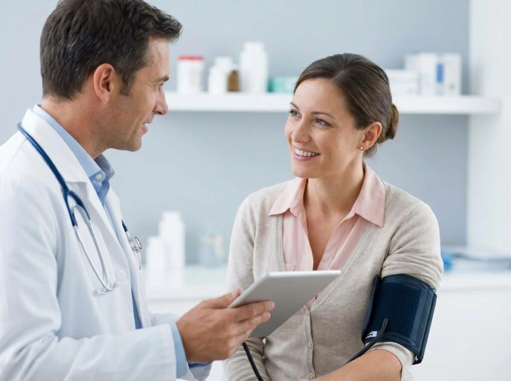 A male physician wearing a white lab coat with a stethoscope around his neck holding a digital tablet and talking with a smiling female patient who has a blood pressure cuff wrapped around her upper arm